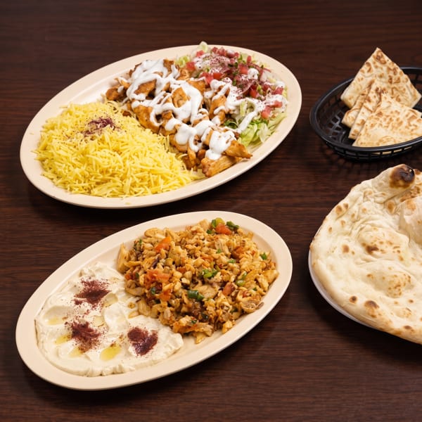 Two plates of Middle Eastern food with chicken, rice, hummus, and pita bread on a wooden table.