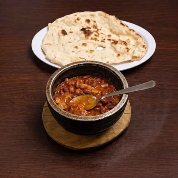 A bowl of kidney bean curry with a spoon, and a plate of naan bread on a wooden table.