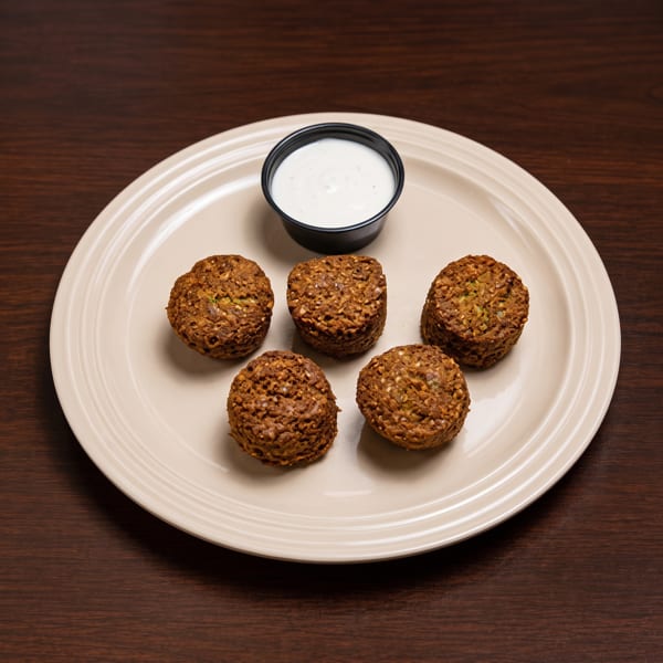 Five crispy brown falafel balls arranged on a beige plate with a side of white dipping sauce.